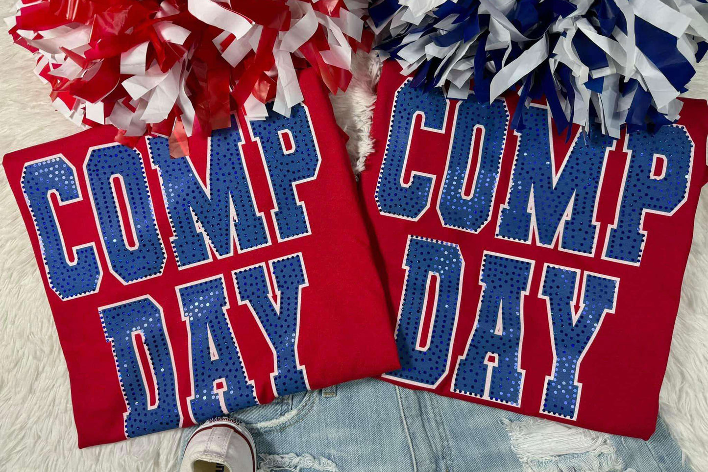 Two red t-shirts laid flat with 'COMP DAY' written in blue and white letters, surrounded by red and white pom-poms and a pair of white sneakers.